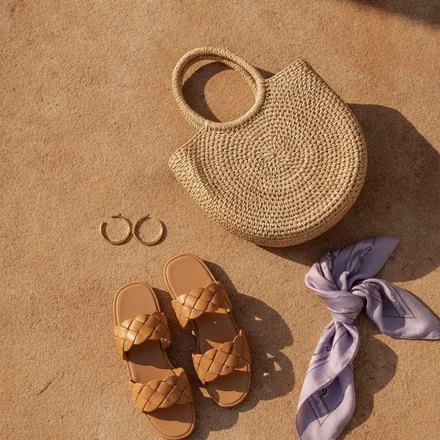 Resort accessories still life: raffia bag, gold hoops, tan sandals, lilac scarf